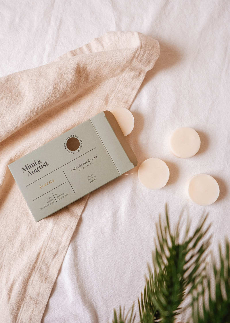 An open box labeled "Soy Wax Melts - Foresta" from Mimi & August rests on a white fabric, displaying three round cream-colored eco-friendly soy wax melts next to it. A sprig of greenery appears in the foreground, promising a long-lasting fragrance that embodies nature's essence.