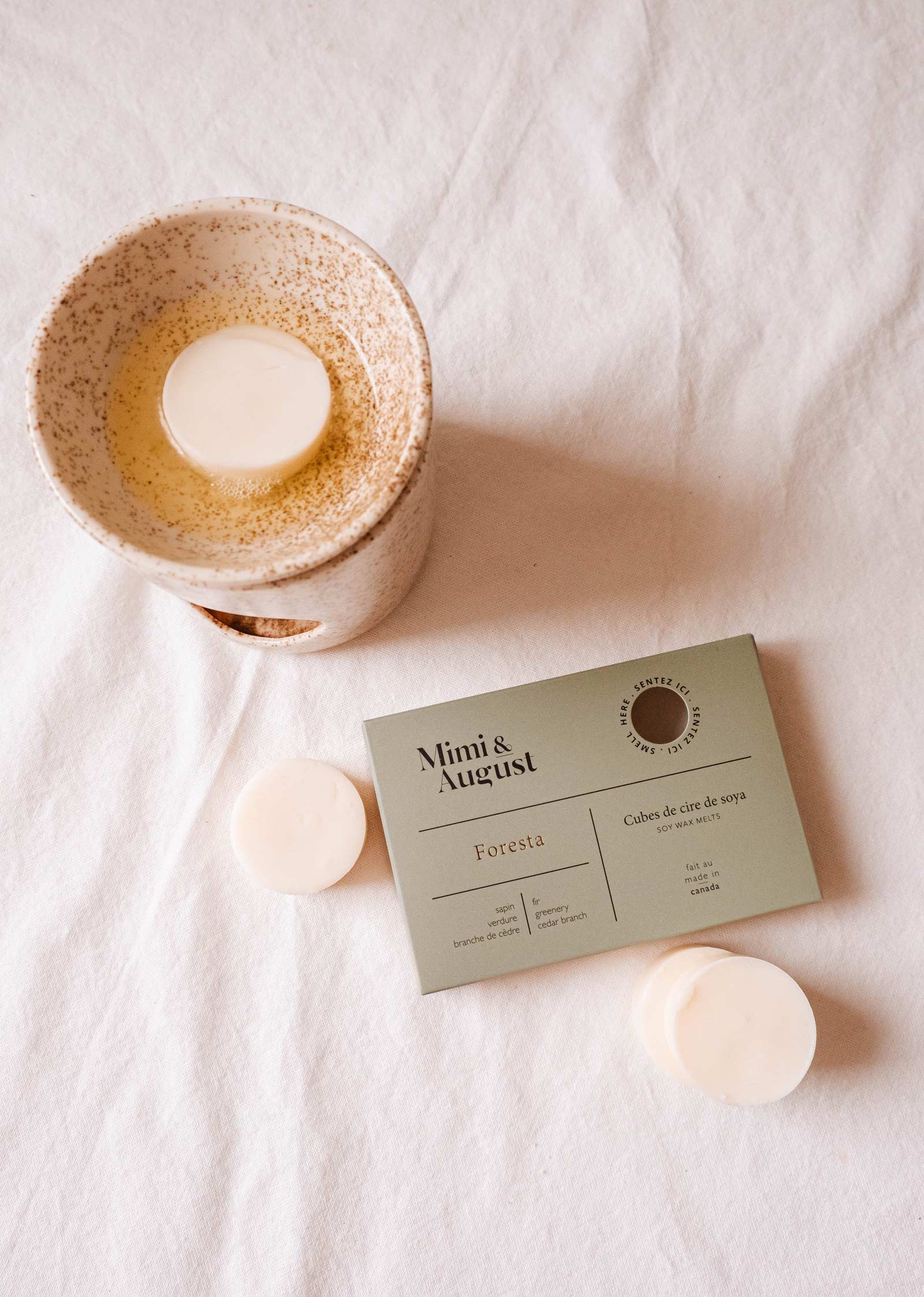 Un bol en céramique contenant une cire fondue repose à côté d'un emballage vert portant la mention "Soy Wax Melts - Foresta" de Mimi & August, accompagné de deux autres cires fondues de soja naturel, le tout sur un fond de tissu blanc. Immergez-vous dans le parfum vivifiant de la forêt de sapins et faites entrer la sérénité de la nature dans votre maison.