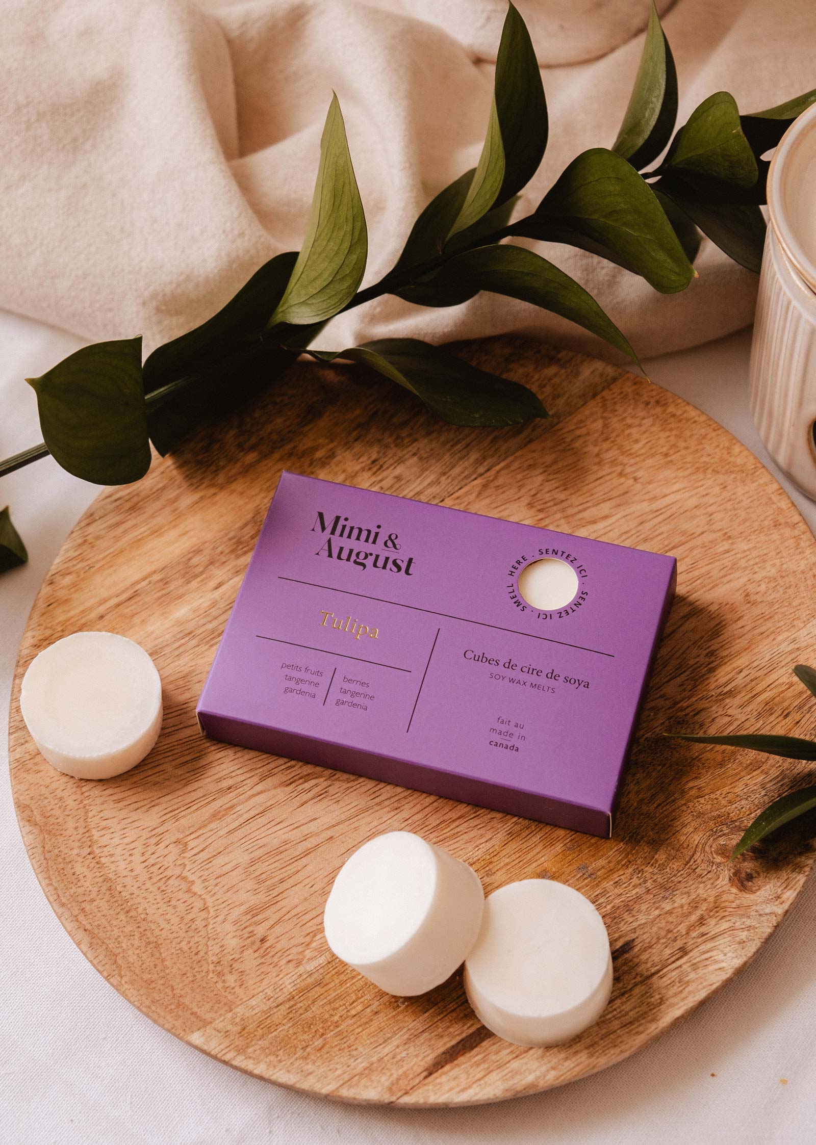 A purple Mimi & August Soy Wax Melts - Tulipa box with natural soy wax melts is shown on a round wooden tray, surrounded by greenery and loose wax melt discs.