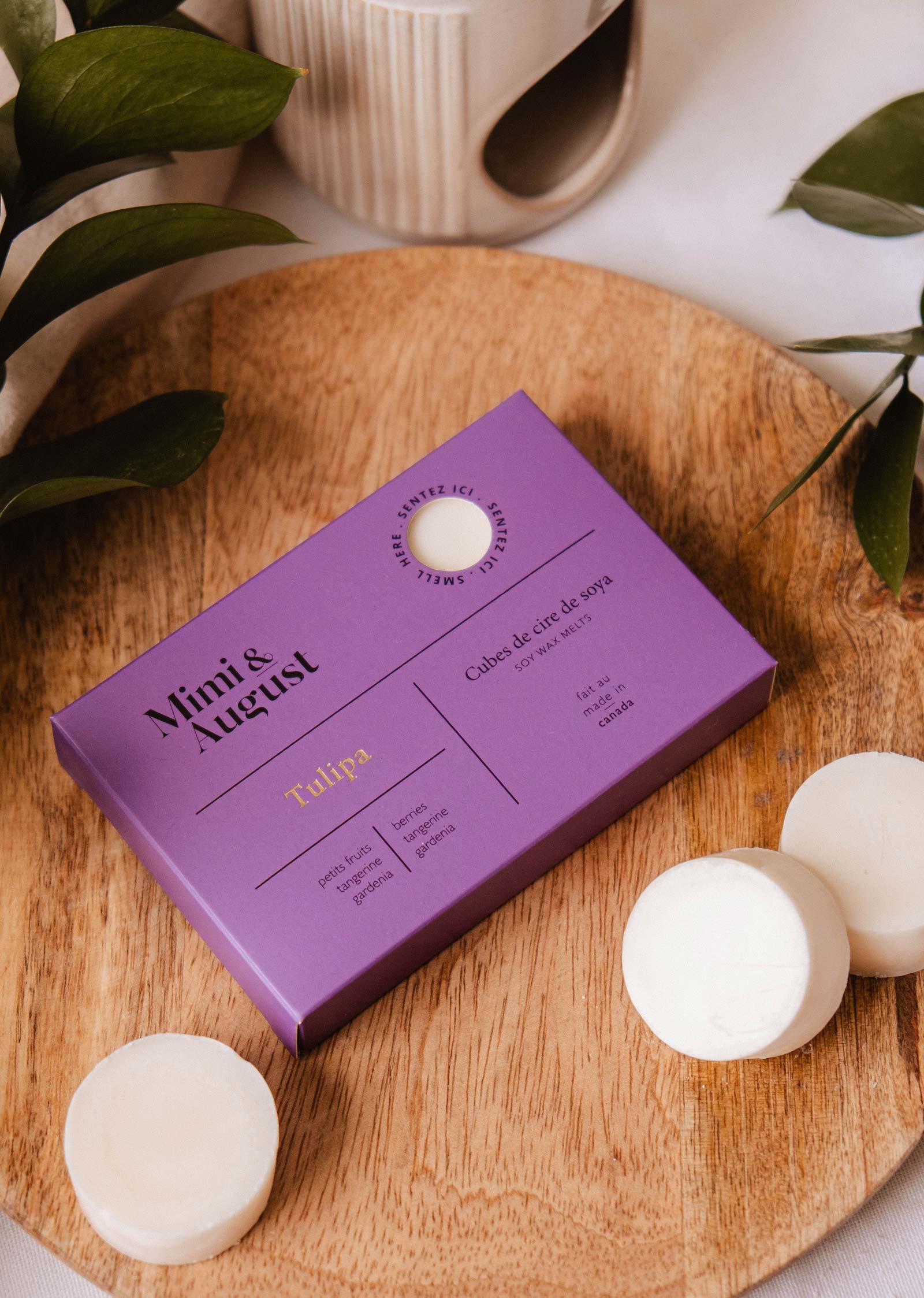 A box of Mimi & August Soy Wax Melts - Tulipa in purple rests on a round wooden tray with three white natural soy wax melts and green leaves nearby.