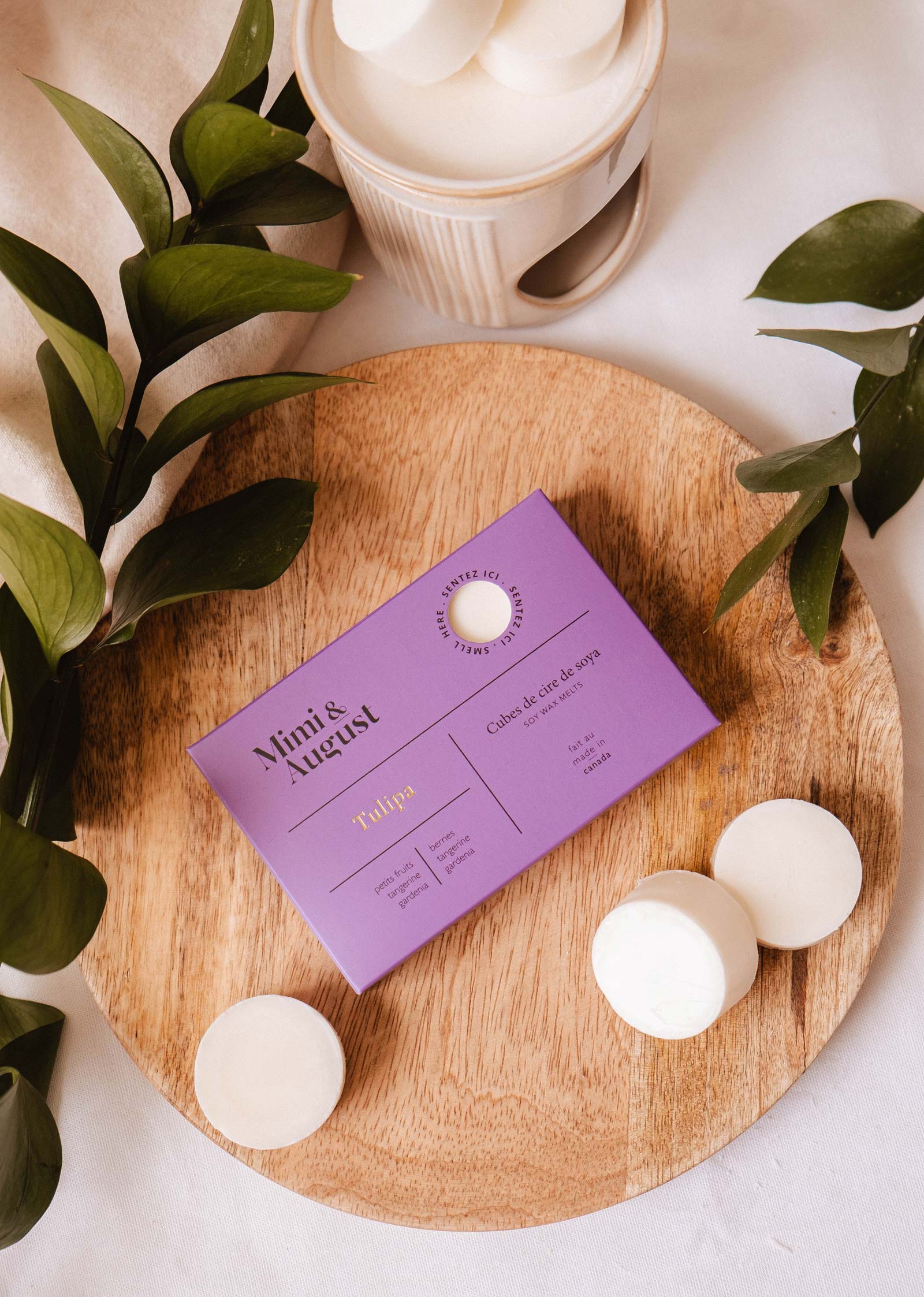 A purple Mimi & August Soy Wax Melts - Tulipa box sits on a wooden tray with round white soaps, green leaves, and a white container—emphasizing its natural soy wax and long-lasting fragrance.