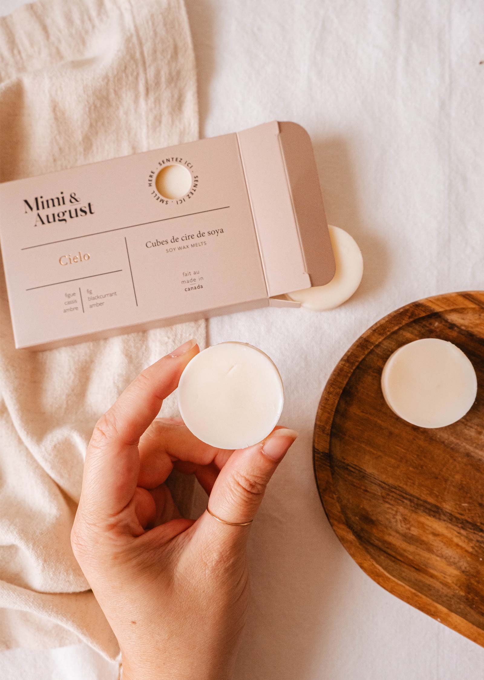 A hand holds a Soy Wax Melts - Cielo, famous for its long-lasting fragrance, near an open "Mimi & August" box on a wooden tray with a cream fabric background.