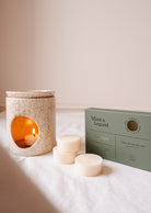 A candle warmer with a lit tealight, accompanied by three cream-colored Soy Wax Melts - Eucalipto from Mimi & August, presented in a green box, provides a soothing natural fragrance on a white surface.