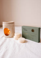 A speckled ceramic candle warmer with a lit candle is positioned beside a box marked "Soy Wax Melts - Eucalipto" by Mimi & August. Three eucalyptus soy wax melts are arranged in front of the box on a white surface, providing a natural fragrance that soothes the senses.