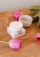 A Candle Refill - Tulipa by Mimi & August and a pink tulip sit on a wooden surface beside a fluted holder, a small bowl, and a box labeled "Mimi & August.