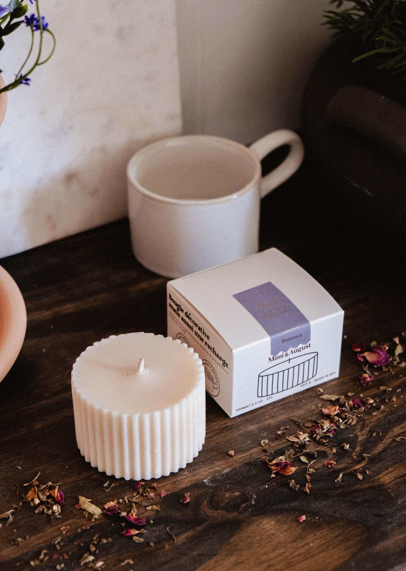 A Mimi & August Candle Refill - Botanica, a box labeled "Mind's Arrest," and a white mug are displayed on a wooden surface with dried flower petals scattered around.