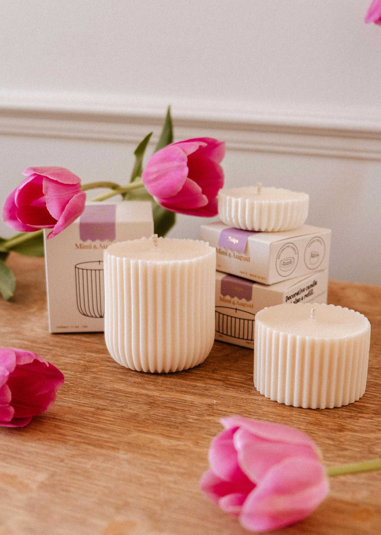 Three Mimi & August Candle Refill - Tulipa soy wax candles of varying sizes are shown on wood, with pink tulips and boxes in the background, adding a touch of tulip scent to the scene.