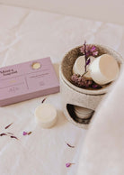 A beige wax melt burner with eco-friendly Mimi & August Soy Wax Melts - Lilac and dried flowers sits by a matching lilac box, filling the air with long-lasting fragrance atop a white fabric surface.