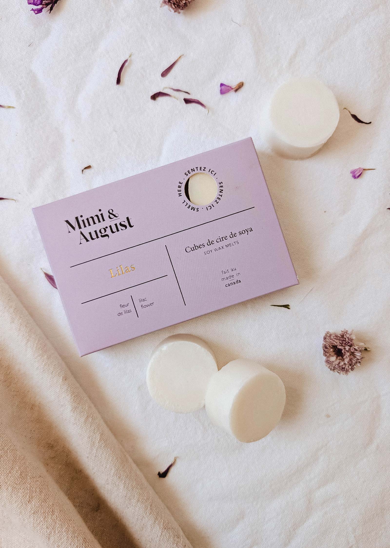 A lavender box labeled "Mimi & August Soy Wax Melts - Lilac" rests on light fabric with three white melts and dried flower petals, offering a long-lasting lilac fragrance for your space.