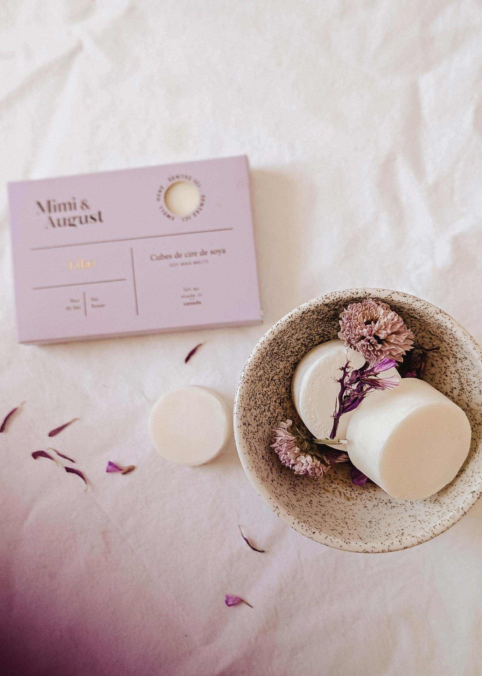 A speckled bowl with two Mimi & August Soy Wax Melts - Lilac and dried flowers sits beside a lavender box on a white surface, providing an eco-friendly, long-lasting fragrance for your space.