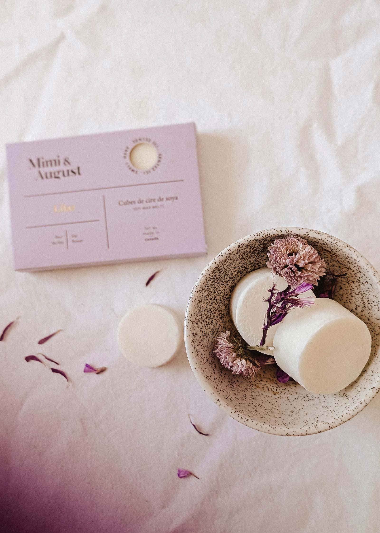 A speckled bowl with two Mimi & August Soy Wax Melts - Lilac and dried flowers sits beside a lavender box on a white surface, providing an eco-friendly, long-lasting fragrance for your space.