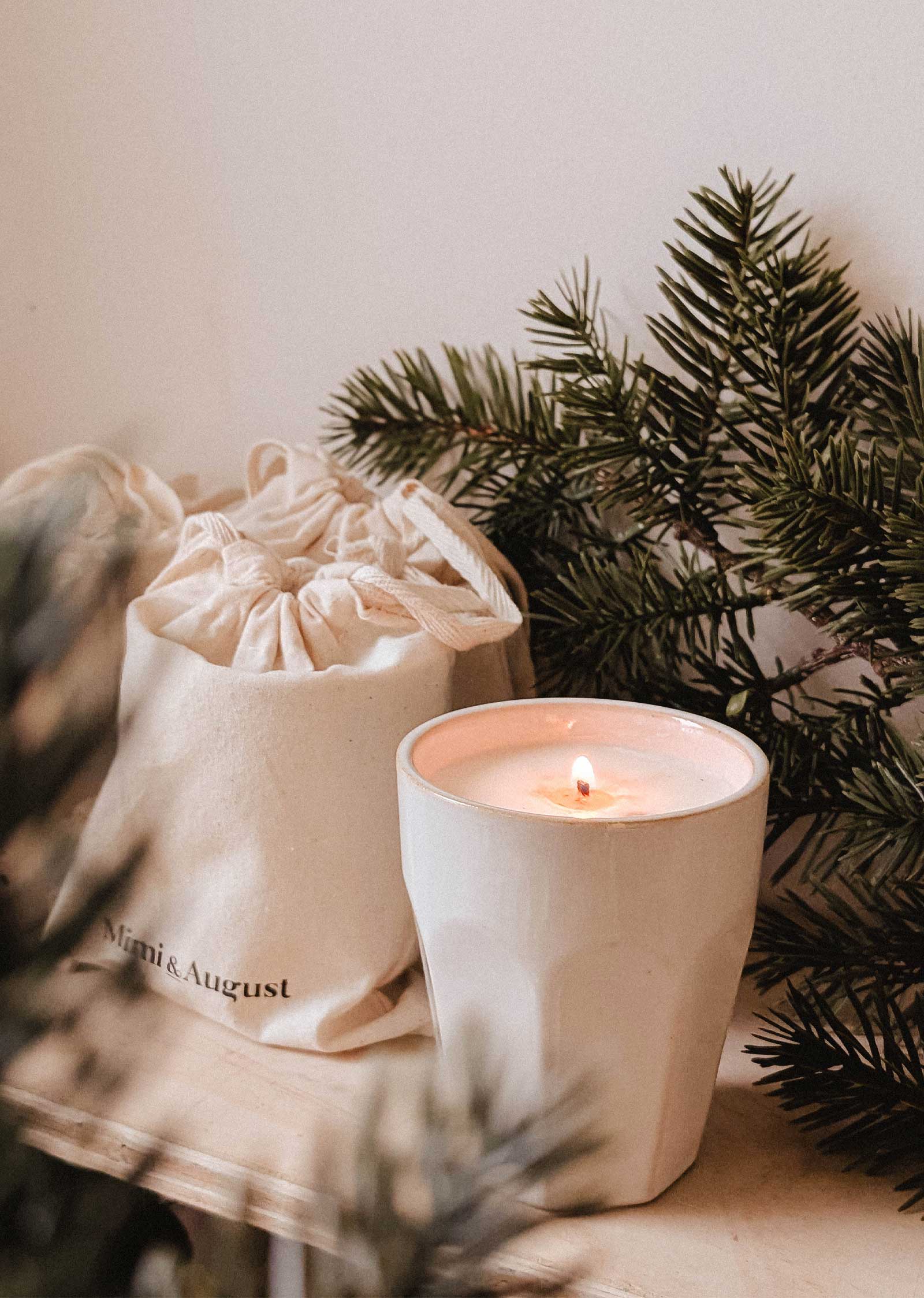 A lit white Special Edition Candle by Mimi & August in a ceramic holder sits beside a drawstring pouch bearing the brand’s name, with pine branches in the background.