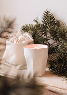 A lit Mimi & August Special Edition Candle in a ceramic holder sits beside a cloth bag labeled with the brand, with pine branches in the background.