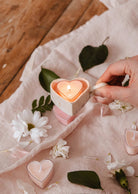 A hand holds a Mimi & August Speckled Heart Tealight Holder with a heart-shaped candle, surrounded by white flowers and more heart-shaped tealights on soft pink fabric.