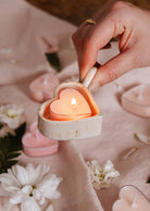 A hand holds a Mimi & August Speckled Heart Tealight Holder with a glowing candle, surrounded by pink heart-shaped tealights and white flowers on a soft fabric surface.
