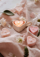 Mimi & August Speckled Heart Tealight Holder displays a glowing heart-shaped tealight, arranged on a soft pink cloth with scattered white flowers and green leaves.
