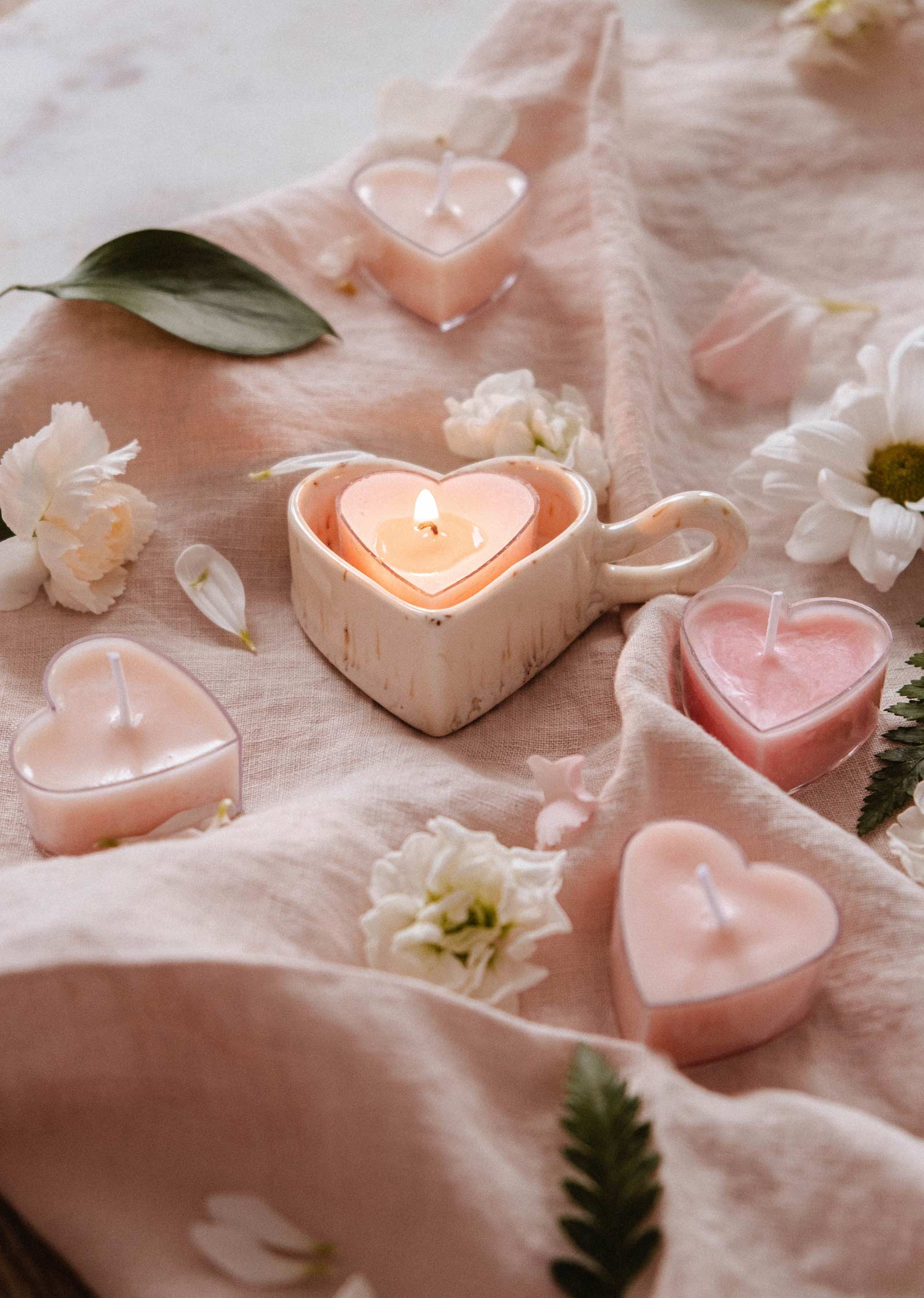 Mimi & August Speckled Heart Tealight Holder displays a glowing heart-shaped tealight, arranged on a soft pink cloth with scattered white flowers and green leaves.