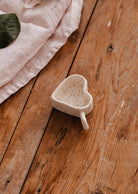 The Mimi & August Speckled Heart Tealight Holder, a small ceramic piece with a handle, rests on a wooden surface beside pale pink fabric.