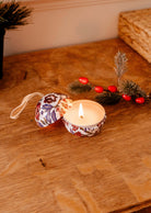 A glowing Spiced Cranberries - Christmas Ornament Candle by Mimi & August in a decorative floral tin on a wooden table, accompanied by a sprig with red berries and elegant ornaments.