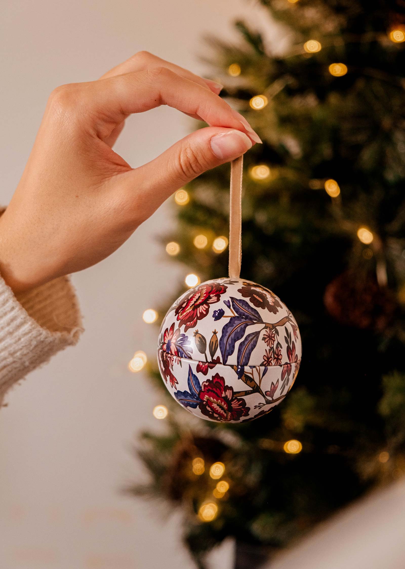 Une main tient délicatement la bougie Spiced Cranberries - Christmas Ornament de Mimi & August, ornée d'un élégant motif floral, scintillant à la lueur d'un arbre de Noël illuminé.