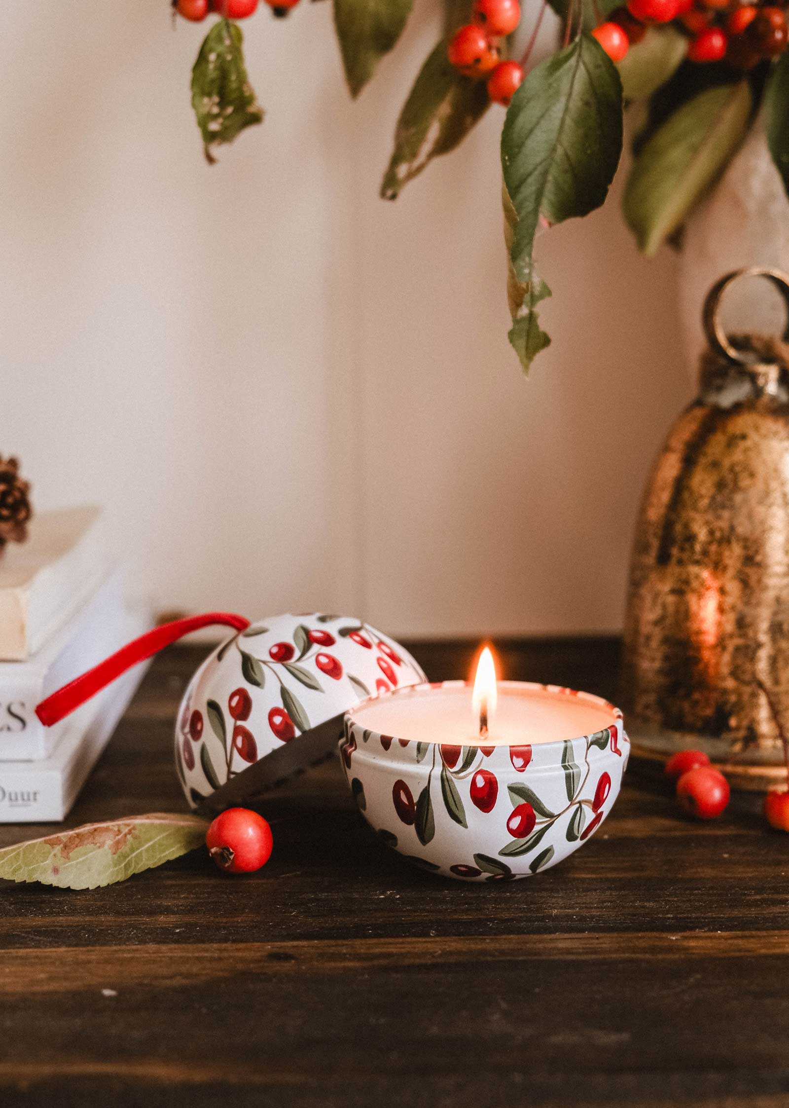 The Mimi & August Spiced Cranberries - Christmas Ornament Candle, in a round berry-patterned container with the lid off, fills the air with a spiced cranberry aroma on a wooden surface near red berries, books, and a brass bell.