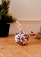 The Spiced Cranberries - Christmas Ornament Candle by Mimi & August, featuring a floral pattern and ribbon loop, sits elegantly on a wooden surface amid greenery and vibrant red berries that evoke the festive spirit in the background.