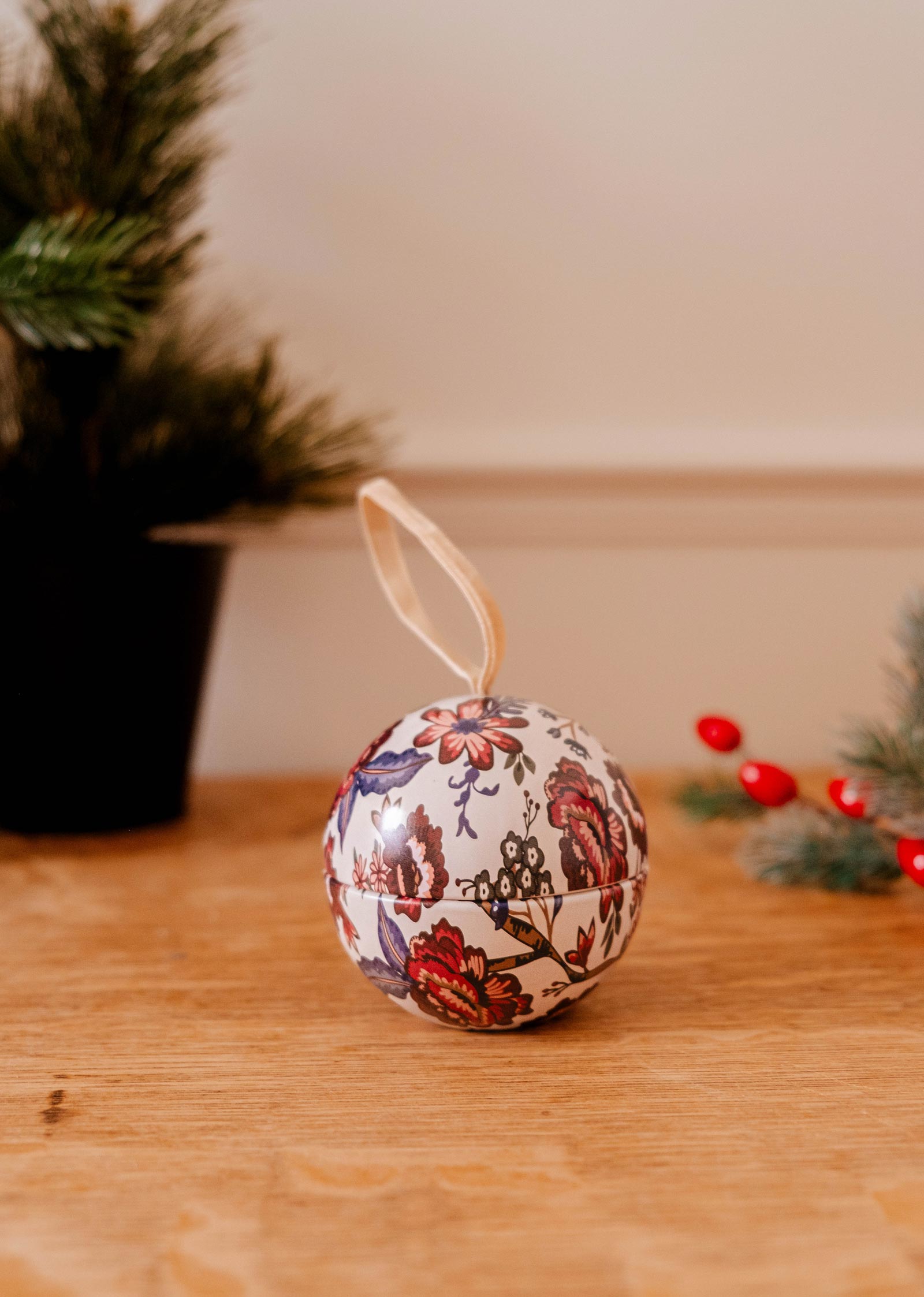 La bougie "Spiced Cranberries - Christmas Ornament" de Mimi & August, ornée d'un motif floral et d'une boucle de ruban, est élégamment posée sur une surface en bois au milieu de verdure et de baies rouges vibrantes qui évoquent l'esprit de fête en arrière-plan.