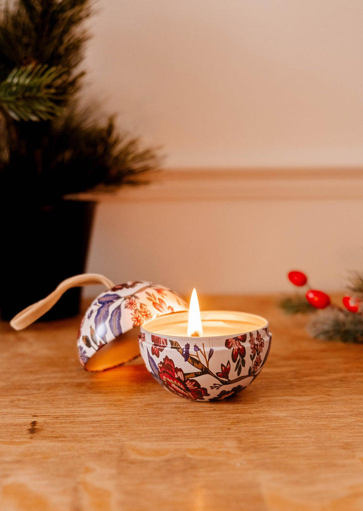 On a wooden surface, the Spiced Cranberries - Christmas Ornament Candle by Mimi & August flickers inside a decorative holder, surrounded by elegant ornaments and greenery, casting a warm glow.