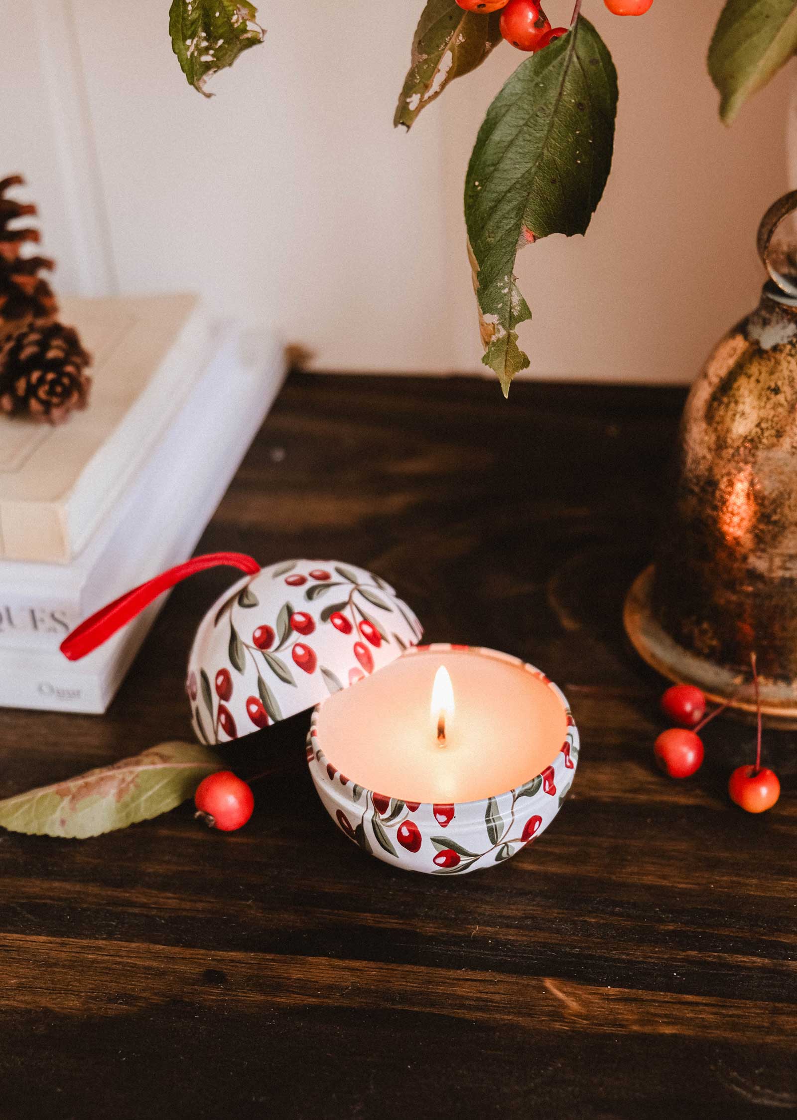 The Mimi & August Spiced Cranberries - Christmas Ornament Candle, featuring a cherry pattern and festive scent in a round container, sits on a wooden table beside a closed book, leaf, and red berries.