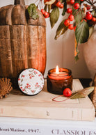 The Mimi & August Spiced Cranberry - 2 oz candle, in a berry-themed tin, is arranged with pinecones and red berries on stacked books for a cozy, festive display perfect for autumn or the holidays.