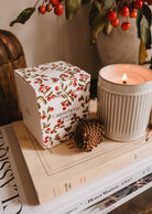 A lit Mimi & August Spiced Cranberry - Christmas Candle, a pinecone, and a cranberry-themed box rest on two stacked art books beside a branch with red berries.