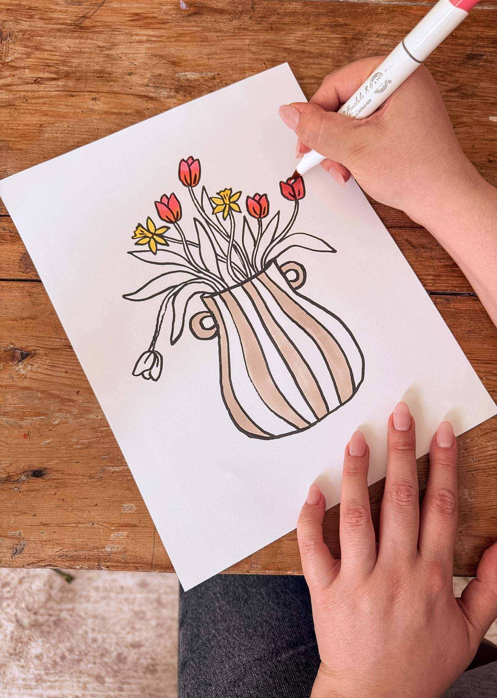A person colors a Mimi & August Spring Bouquet Coloring Sheet, which shows flowers in a striped vase on white paper, using a red marker at a wooden table—a free sheet perfect for creative fun.