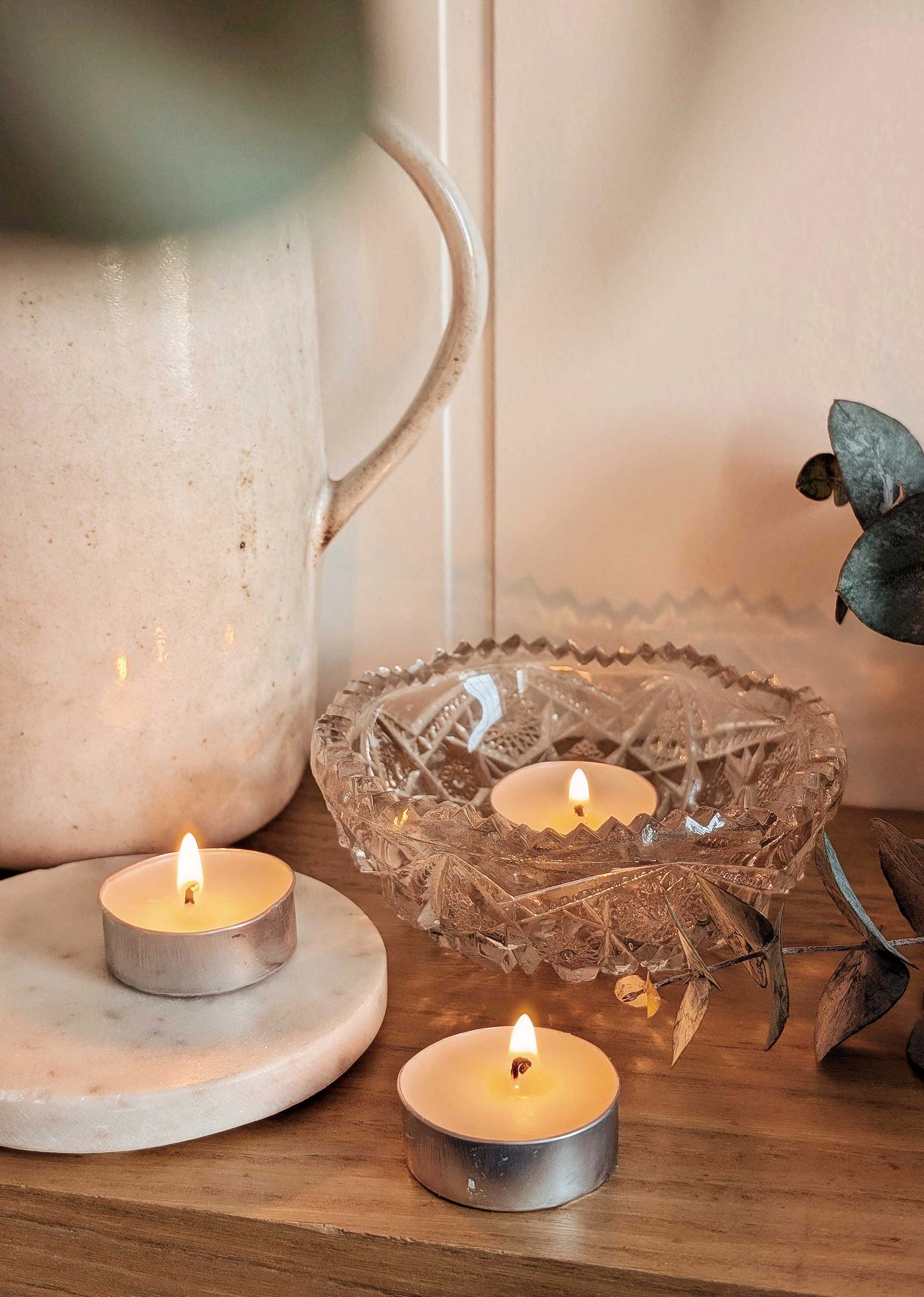 Bougies de thé allumées sur une surface en bois, accompagnées d'un bol en verre décoratif, d'un pichet en céramique et de feuilles d'eucalyptus provenant du kit de découverte exclusif de Mimi & August.