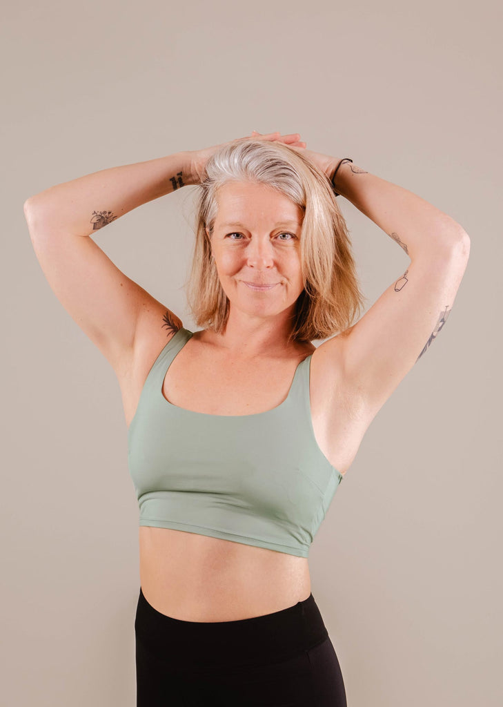 A middle-aged woman with grey hair smiling, wearing a green Mimi & August Tahiti Agave Bralette Bikini Top and black leggings, posing with hands behind head against a beige background.