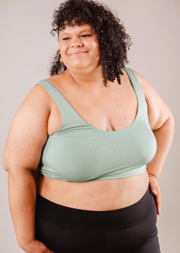 A confident plus-size woman with curly hair, wearing a Mimi & August Tahiti Agave Bralette Bikini Top and black leggings, stands with hands on hips against a neutral background.