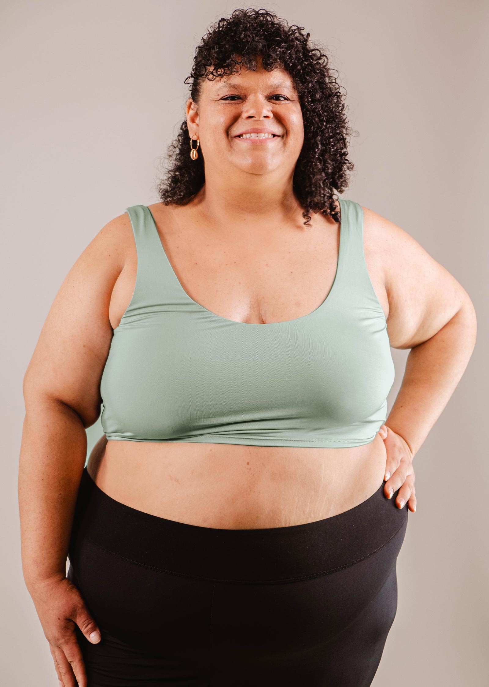 A smiling woman with curly hair, wearing a Mimi & August Tahiti Agave Bralette Bikini Top and black leggings, standing against a neutral background.