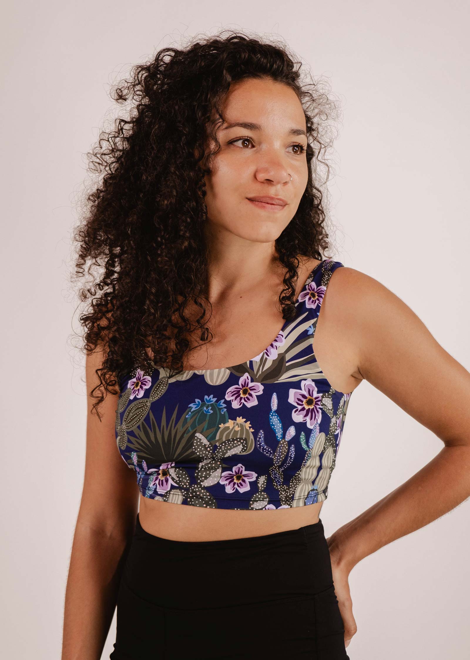 Une personne aux cheveux bouclés, vêtue d'un haut de bikini Mimi & August Tahiti Jardin de Nuit Bralette à motifs bleus et floraux et d'un bas de bikini noir, se tient debout sur un fond uni.