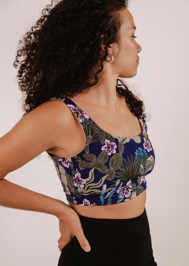 A woman with curly hair wearing a Mimi & August Tahiti Jardin de Nuit Bralette Bikini Top and black pants stands with one hand on her hip, looking to the side.