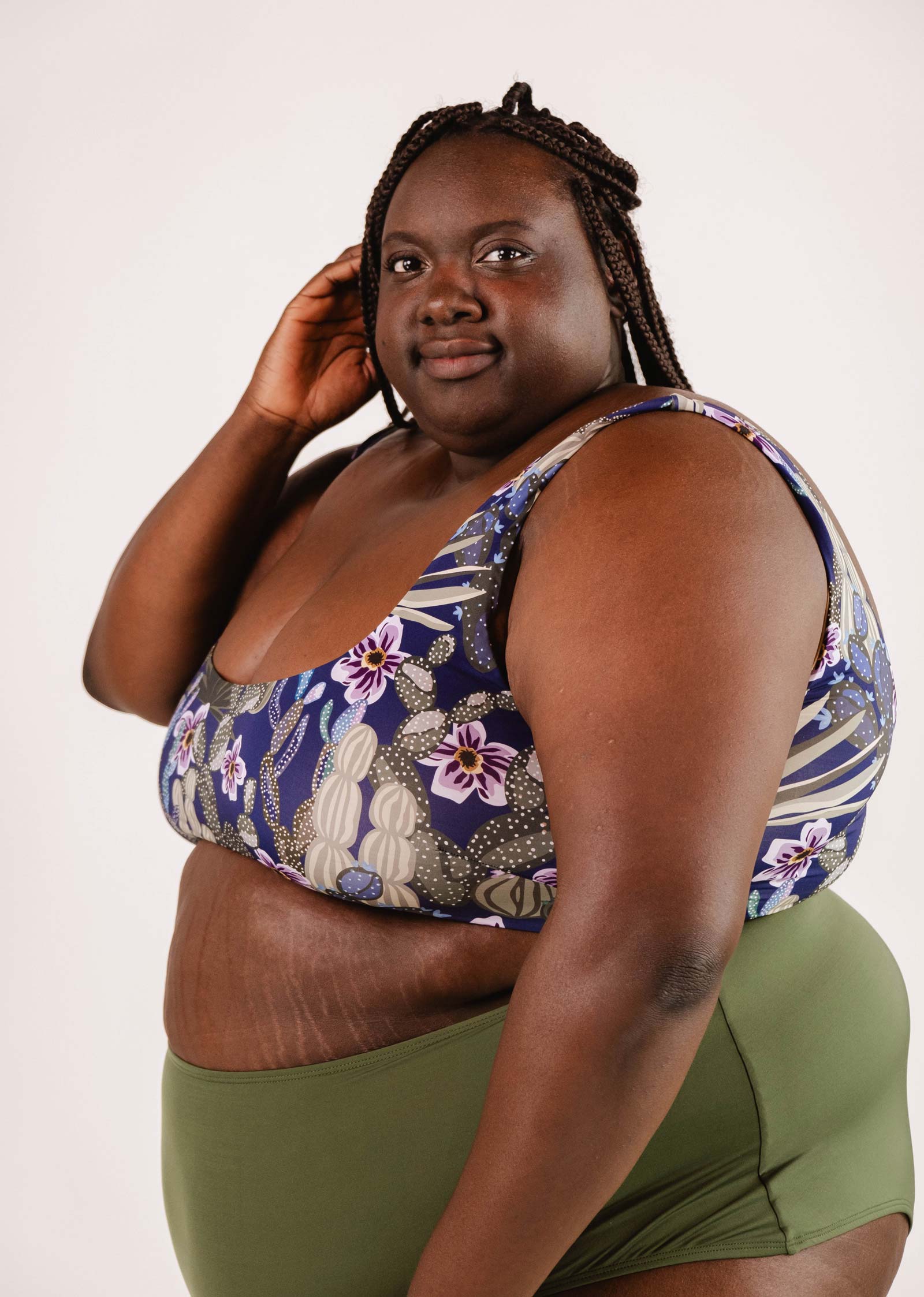 A woman with braided hair stands confidently in a blue Mimi & August Tahiti Jardin de Nuit Bralette Bikini Top and green bottoms, looking at the camera with a neutral expression.