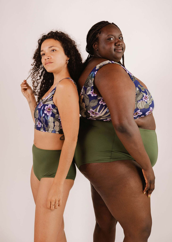 Two women stand back-to-back, adorned in vibrant floral print tops and green bottoms. The woman on the left, with curly hair, wears a Tahiti Jardin de Nuit Bralette Bikini Top from Mimi & August featuring a tropical print. The woman on the right sports braided hair and exudes confidence in her Tahiti Jardin de Nuit ensemble.