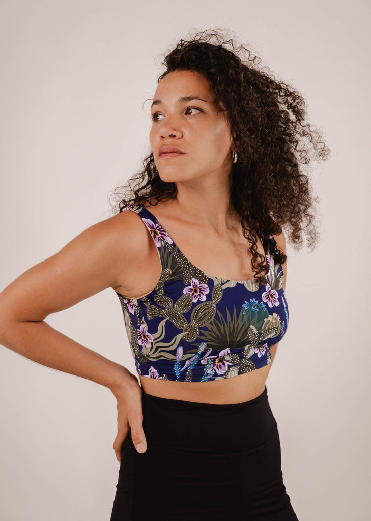 A woman with curly hair wearing a Mimi & August Tahiti Jardin de Nuit Bralette Bikini Top and black leggings stands with her hand on her hip, looking to the side.