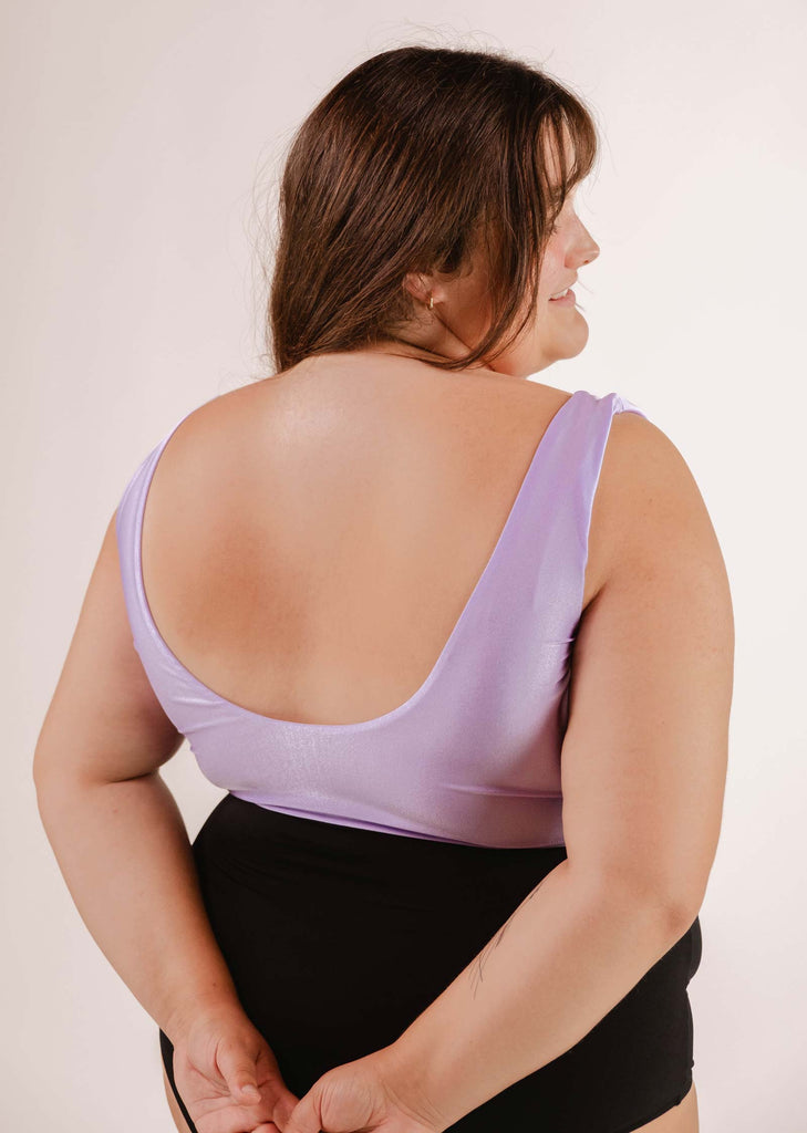 A woman with brown hair wears a vibrant Mimi & August Tahiti Lilac Bralette Bikini Top and black bottoms, facing away from the camera with her hands clasped behind her back.