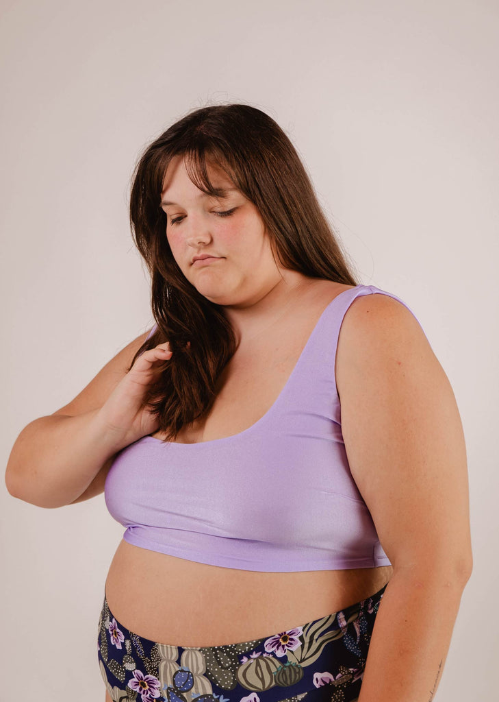 Une personne aux cheveux longs portant un haut de bikini Mimi & August Tahiti Lilas Bralette et un pantalon à motifs, vue en train de toucher ses cheveux sur un fond neutre, exsudant un look de plage sans effort.