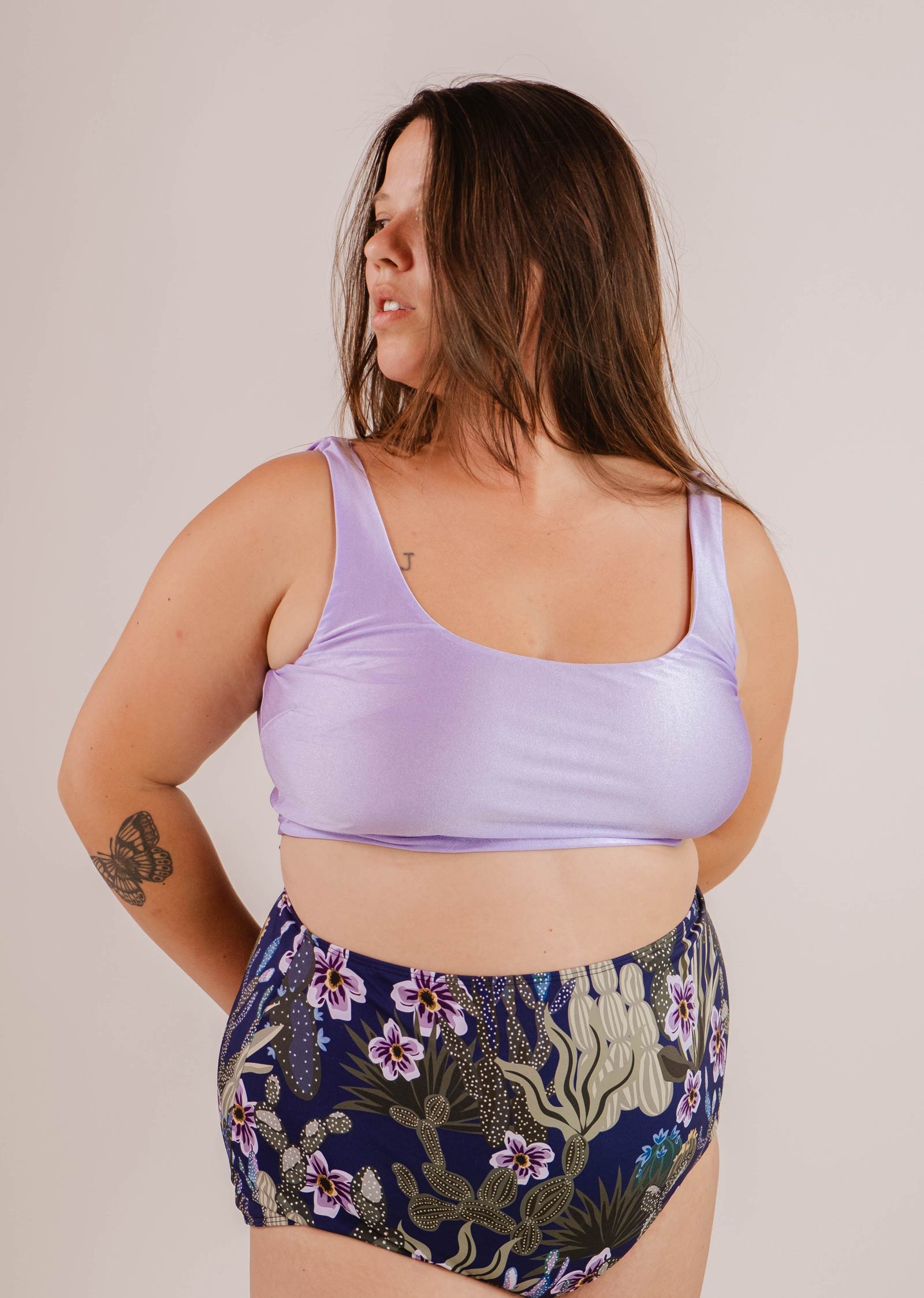 A woman with long brown hair wearing a Mimi & August Tahiti Lilac Bralette Bikini Top and floral high-waisted bikini bottoms stands with one arm bent and looks to the side.