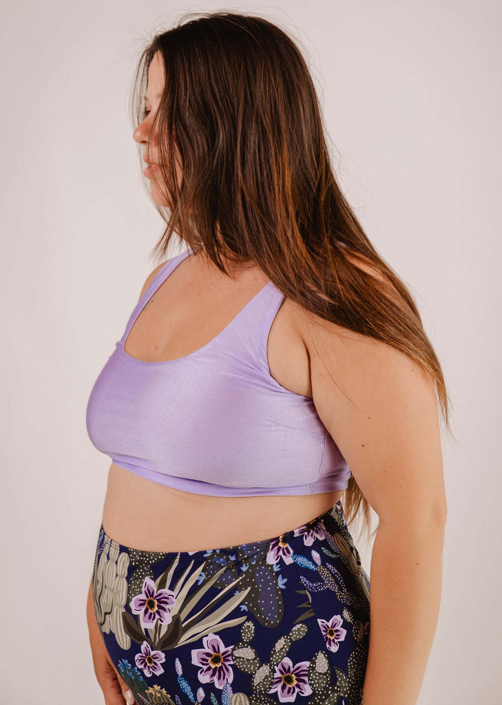 Une personne aux cheveux longs et raides, portant un haut de bikini bralette Tahiti Lilas de Mimi & August et un legging à motifs floraux, debout de profil sur un fond uni.