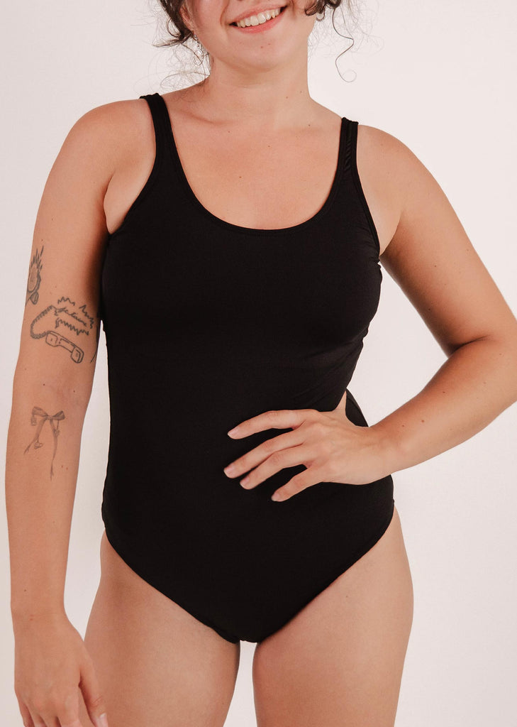 A person wearing a Mimi & August Tamarindo Black One-Piece Swimsuit is smiling with a hand on their hip, ready for beach activities. Visible tattoos adorn their arm.