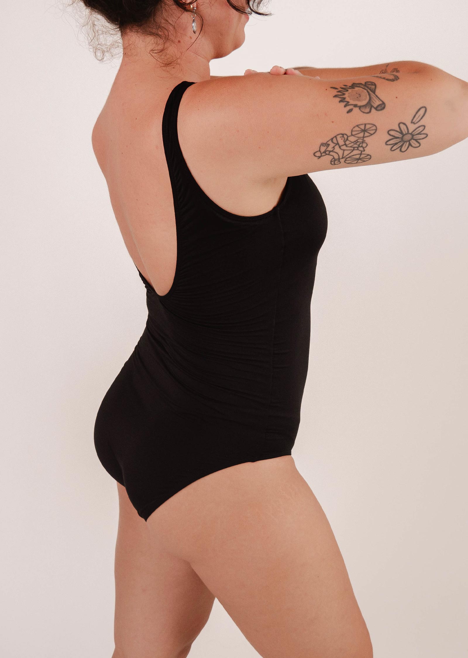 Une personne tatouée sur l'épaule et le bras se tient de côté, vêtue d'un élégant maillot de bain une pièce Tamarindo Black de Mimi & August, idéal pour les activités de plage.