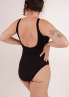 Personne portant un maillot de bain une pièce Tamarindo Black, un classique intemporel de Mimi & August, montré de dos, les mains sur les hanches et les tatouages visibles sur le bras droit. Le maillot de bain offre un bon soutien et met en valeur l'assurance de la personne.