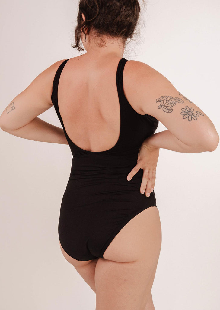 Person wearing a timeless classic Tamarindo Black One-Piece Swimsuit by Mimi & August shown from the back, with hands on hips and visible tattoos on the right arm. The swimsuit offers good support and enhances their confident stance.