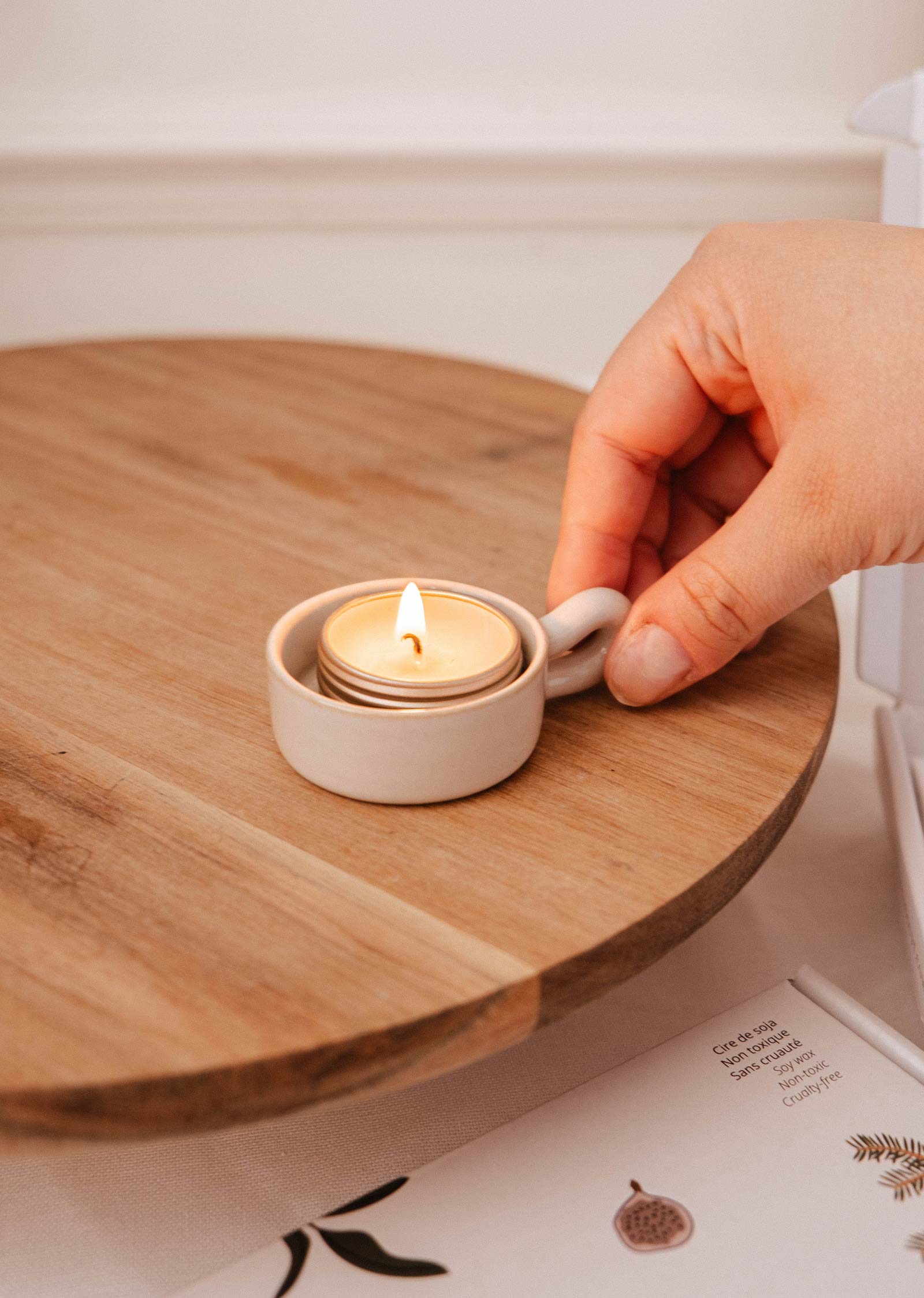 A hand places a lit tealight in the Mimi & August Tealight Holder, featuring a white ceramic base on a round wooden surface—an ideal accent for rustic decor.